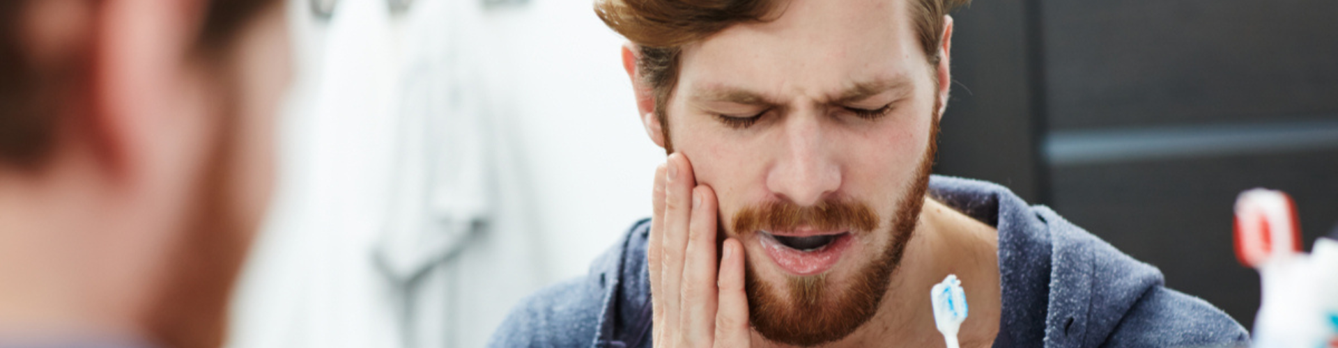 A man pausing while brushing his teeth, holding his cheek in pain from a toothache.